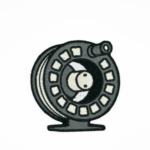 May include: Embroidered patch depicting a grey and black fishing reel. The reel has a circular design with white square accents around the edge. A small handle is visible on the side. The patch is designed for application on clothing or accessories.