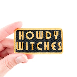 May include: A black iron-on patch with gold embroidered text that reads "Howdy Witches".
