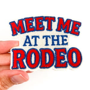 Può includere: Toppa ricamata con la scritta "MEET ME AT THE RODEO" in lettere rosse con contorni blu. La toppa ha uno sfondo bianco ed è tenuta da una mano su uno sfondo bianco.