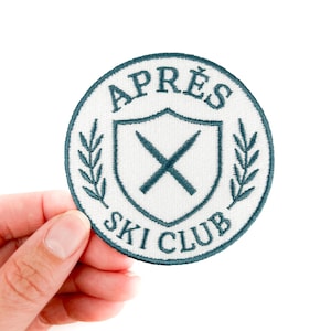 May include: Round embroidered patch with the text "APRES SKI CLUB" in teal. The design includes a shield with crossed skis and laurel leaves. The patch has a white background and is suitable for clothing or accessories.