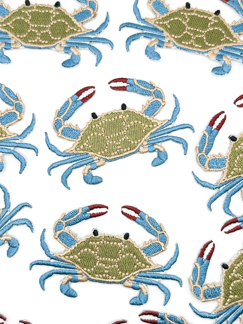 May include: A repeating pattern of embroidered crab patches on a white background. Each crab features a textured olive green shell, bright blue legs and claws, and distinct red tips on the pincers. The embroidery uses visible stitching.