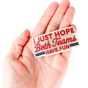 K&ouml;nnte beinhalten: Ein rechteckiger Aufn&auml;her mit dem Text "I JUST HOPE Both Teams HAVE FUN" in roter und wei&szlig;er Schrift. Der Aufn&auml;her hat einen roten Rand und blaue Streifen. Der Aufn&auml;her wird in einer Hand gehalten.