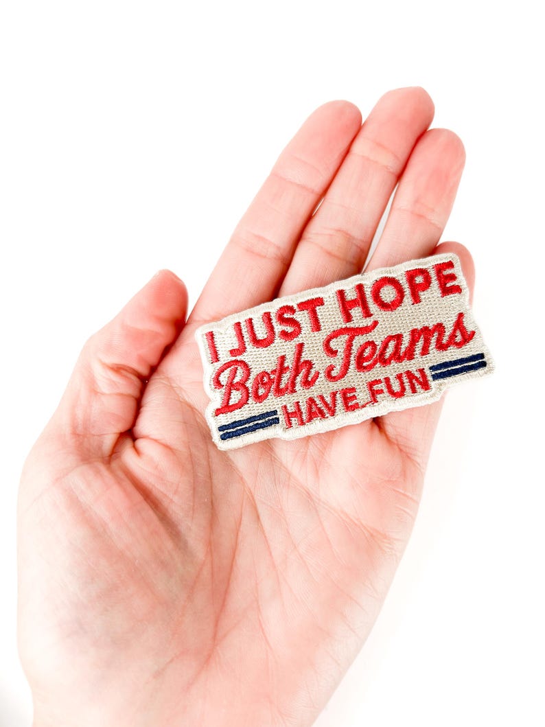 K&ouml;nnte beinhalten: Ein rechteckiger Aufn&auml;her mit dem Text "I JUST HOPE Both Teams HAVE FUN" in roter und wei&szlig;er Schrift. Der Aufn&auml;her hat einen roten Rand und blaue Streifen. Der Aufn&auml;her wird in einer Hand gehalten.