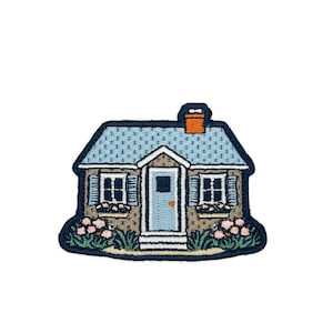 May include: Embroidered patch depicting a quaint cottage with a blue roof, light blue door, and window shutters. The house has a brown facade, a brick chimney, and is surrounded by pink flowers and green foliage.