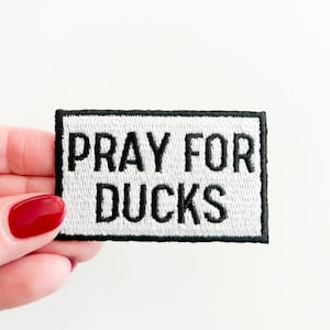 May include: White embroidered patch with black stitching. The patch reads "Pray for Ducks".