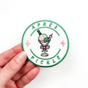 May include: A white embroidered patch with a green border. The patch features a green cocktail glass with a pink drink, a green lime wedge, a cherry, and a bottle of liquor. The text "APRES PICKLE" is written in a green font around the edge of the patch.