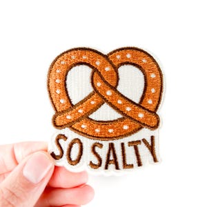 May include: Embroidered patch featuring a pretzel design with brown and white details. The patch has the text "SO SALTY" in brown lettering. The patch is held against a white background.