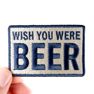 May include: Embroidered patch with the words "WISH YOU WERE BEER" in navy blue thread on a rectangular patch. The patch has a light beige background and a navy blue border. The patch is held by a hand.