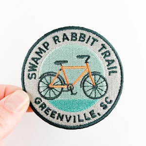 Op de afbeelding: Geborduurde ronde patch met een oranje fietsillustratie. De patch heeft de tekst "SWAMP RABBIT TRAIL GREENVILLE, SC" in donkergroen. De achtergrond is lichtblauw en turquoise.