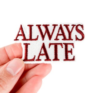 May include: White rectangular patch with the embroidered words "ALWAYS LATE" in burgundy. The patch is held by a hand against a white background. The patch is a textile item.
