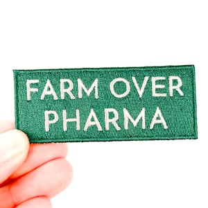 May include: A rectangular green patch with white embroidered text that reads "FARM OVER PHARMA." The patch is held in a hand against a white background. The patch is a textile item.