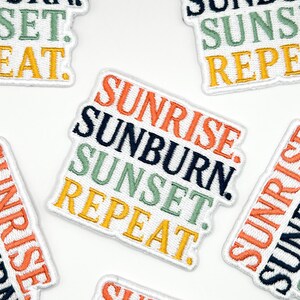 以下が含まれることがあります： SUNRISE、SUNBURN、SUNSET、REPEATという文字が刺繍された5つのワッペン。各単語はオレンジ、ダークブルー、セージグリーン、イエローの異なる色で刺繍されています。ワッペンは白い背景で、白い刺繍の縁取りが施されています。