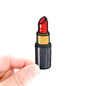 Puede incluir: Parche bordado de un labial rojo con un tubo negro y dorado. El labial tiene un color rojo brillante y está sobre un fondo blanco. El parche es un accesorio de moda.