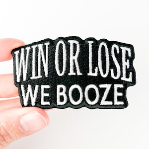 以下が含まれることがあります： 白のスタイリッシュな文字で「WIN OR LOSE WE BOOZE」と書かれた黒色の刺繍パッチ。パッチは波状の縁を持つ黒い縁取りがあります。白い背景に対して、手でパッチが持たれています。