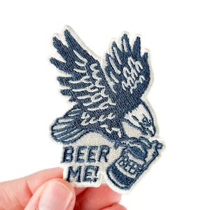 May include: Embroidered patch featuring a navy blue eagle carrying a beer bottle. The patch has the words "BEER ME!" in bold letters. The eagle and beer bottle are detailed with white stitching.