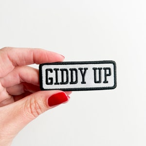 May include: A rectangular patch with the words "GIDDY UP" embroidered in black on a white background. The patch has a black border. The patch is held by a person with red nail polish.