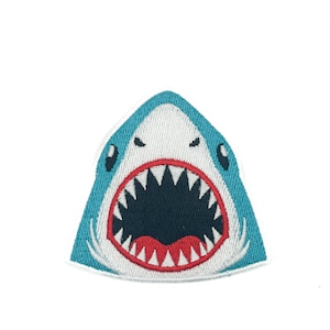 Op de afbeelding: Een geborduurde patch in de vorm van een haaienkop met een wijd openstaande bek. De haai heeft een blauwgroene rand, witte huid, een donker marineblauwe binnenkant van de bek, scherpe witte tanden en een rode tong, gedetailleerd gestikt.