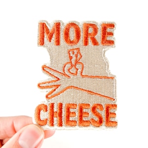 May include: Embroidered patch with the words "MORE & CHEESE" in orange thread. The patch has a light beige background with an orange cheese and scissors graphic. The patch is held by a hand against a white background.