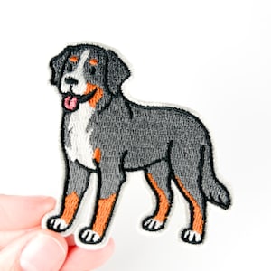 Könnte beinhalten: Gestickter Aufnäher mit einem Berner Sennenhund. Der Hund wird in einer stehenden Pose mit einem Farbschema aus Schwarz, Grau, Weiß und Orange dargestellt. Der Aufnäher hat einen weißen Rand und ist zum Aufbügeln oder Aufnähen auf Stoff vorgesehen.