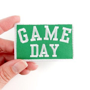 Puede incluir: Un parche de tela verde con texto bordado en blanco que dice "GAME DAY".
