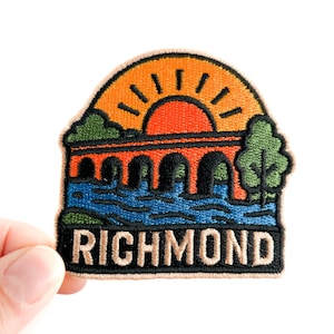 Puede incluir: Parche bordado con un diseño de paisaje que representa un puente sobre un río, un sol naciente y la palabra "RICHMOND". El parche es de color naranja, azul, verde y negro. Es un recuerdo.