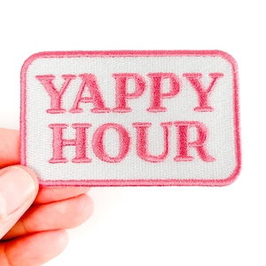 Könnte beinhalten: Ein rechteckiger Aufnäher mit den Worten "YAPPY HOUR" in Rosa auf weißem Grund gestickt. Der Aufnäher hat einen rosa Rand und wird von einer Hand vor weißem Hintergrund gehalten.