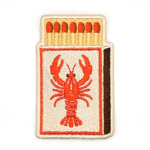 May include: An embroidered patch shaped like a matchbox with a cream background and a dark brown side strip. The center features a bright orange lobster graphic. The top section displays a row of six matches with tan stems and orange tips.
