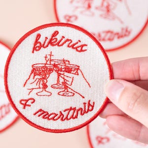 May include: Embroidered patch with a white background and a red border. The patch features the words "bikinis & martinis" in red script, along with a red illustration of three martini glasses being clinked together. The patch is round.