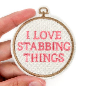 Op de afbeelding: Geborduurde patch met de tekst "I LOVE STABBING THINGS" in roze, geplaatst in een witte cirkel met een bruine rand. De patch heeft een lus aan de bovenkant en wordt door een hand vastgehouden.