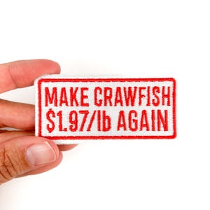 May include: A rectangular white patch with a red border and red embroidered text. The text reads "MAKE CRAWFISH $1.97/lb AGAIN". The patch is held by a hand against a white background.