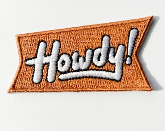Könnte beinhalten: Ein brauner Filz-Patch mit weiß gesticktem Schriftzug "Howdy!"