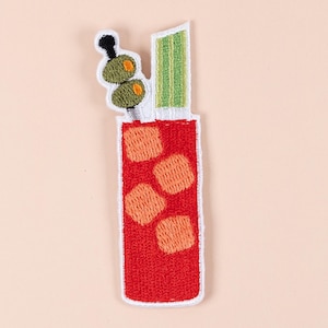 以下が含まれることがあります： ブラッディマリーカクテルの刺繍パッチ。赤い飲み物には、オレンジ色の氷、セロリの茎、楊枝に刺したオリーブが飾られています。パッチは白い縁取りで、明るいピーチ色の背景に配置されています。