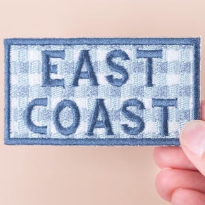 May include: A rectangular embroidered patch featuring the text "EAST COAST" in navy blue. The background has a blue and white gingham pattern, with a navy blue border. The patch is held by a hand.
