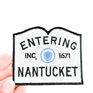 Könnte beinhalten: Ein weißer, bestickter Aufnäher mit schwarzem Rand. Der Aufnäher zeigt die schwarzen Worte „ENTERING NANTUCKET“ sowie „INC. 1671“ und ein hellblaues Emblem.