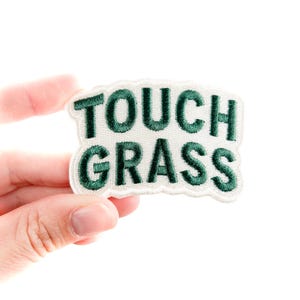 以下が含まれることがあります： 緑色の「TOUCH GRASS」の文字が刺繍された白いパッチ。パッチは白い縁取りがあり、手で持たれています。テキストは太字のサンセリフ体です。背景は白です。