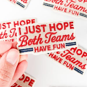 Può includere: Toppe ricamate con la scritta "I JUST HOPE Both Teams HAVE FUN" in lettere rosse su sfondo crema. Ogni toppa ha due strisce blu e un bordo rosso. Le toppe sono sparse su una superficie bianca.