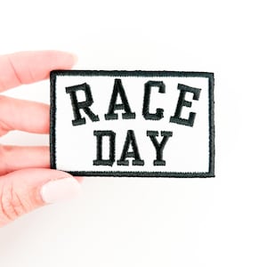 May include: White fabric patch with black embroidered text that reads "RACE DAY" surrounded by a black border.
