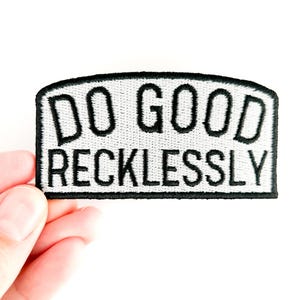 May include: A rectangular patch with a black border and the words "DO GOOD RECKLESSLY" embroidered in black on a white background. The patch is held by a hand against a white backdrop.