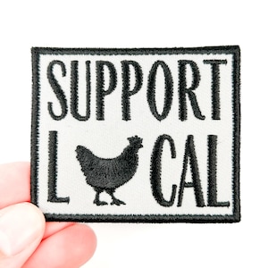 May include: A rectangular patch with a black border and the words "SUPPORT LOCAL" in bold, black capital letters. A black silhouette of a chicken is centered below the word "SUPPORT". The patch is held by a hand.