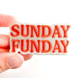 May include: Embroidered patch with the words "SUNDAY FUNDAY" in bright orange letters. The text is stitched onto a light gray background with a white border. The patch is held against a white background.