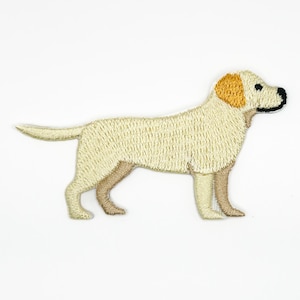 May include: An embroidered patch in the shape of a standing yellow Labrador retriever dog. The dog has light cream-colored fur, a darker golden-yellow ear, and a black nose. The embroidery features detailed stitching to represent the dog's coat.