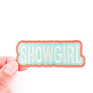 Może przedstawiać: Haftowana naszywka z napisem "SHOWGIRL" w kolorze białym, obramowana jasnoniebieskim i obramowana kolorem koralowym. Naszywka jest prostokątna z zaokrąglonymi rogami.