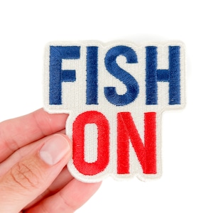 May include: Embroidered patch with the words "FISH ON" in blue and red. The patch has a white border and is held by a hand against a white background. The patch is a fun accessory for fishing enthusiasts.