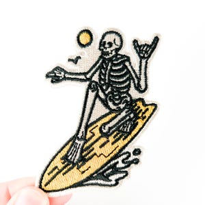 Puede incluir: Parche bordado con un esqueleto surfeando sobre una tabla de surf amarilla. El esqueleto es blanco y negro, con un sol y pájaros en el fondo. El parche está sujeto por una mano sobre un fondo blanco.