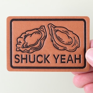 Puede incluir: Un parche rectangular de cuero marrón con un borde negro. El parche presenta dos ilustraciones de ostras y las palabras "SHUCK YEAH" en letras negras. El parche está sostenido por una mano.
