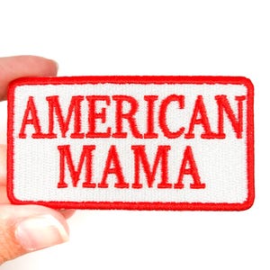 Op de afbeelding: Rechthoekige geborduurde patch met de tekst "AMERICAN MAMA" in rode hoofdletters op een witte achtergrond. De patch heeft een rode rand. De patch wordt door een hand vastgehouden.