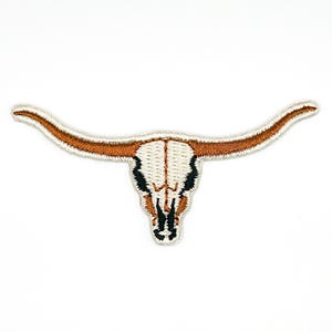 Könnte beinhalten: Ein gestickter Aufnäher, der einen Longhorn-Stierschädel darstellt. Der Schädel ist weiß mit schwarzen Akzenten um die Augenhöhlen und die Nasenhöhle, und die langen, geschwungenen Hörner sind in strukturiertem, rostorangem Garn mit weißem Rand.