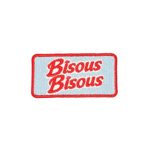 May include: A rectangular patch with a light blue background and a red border. The patch features the words "Bisous Bisous" in red, cursive lettering. The patch is likely an embroidered item.