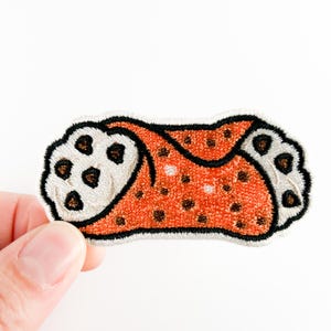 May include: Embroidered patch in the shape of a cannoli. The patch features an orange body with brown speckles and white ends with brown spots. The patch is outlined in black.