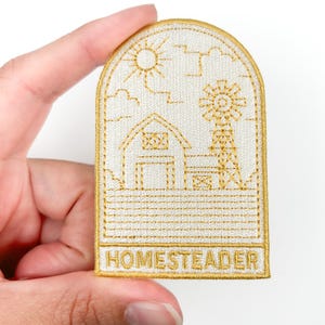 May include: Embroidered patch with a farm scene featuring a barn, windmill, sun, and clouds, outlined in gold on a cream background. The word "HOMESTEADER" is at the bottom. The patch is held by a hand.